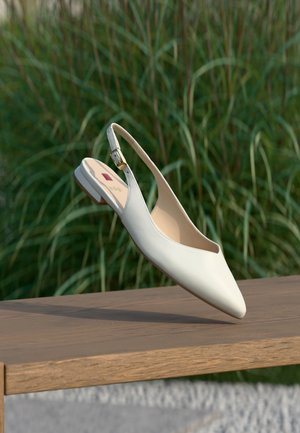 Chaussure plate blanche en cuir avec bride arrière, bout pointu et boucle dorée, présentée en suspension au-dessus d'une surface en bois avec des plantes vertes en arrière-plan.