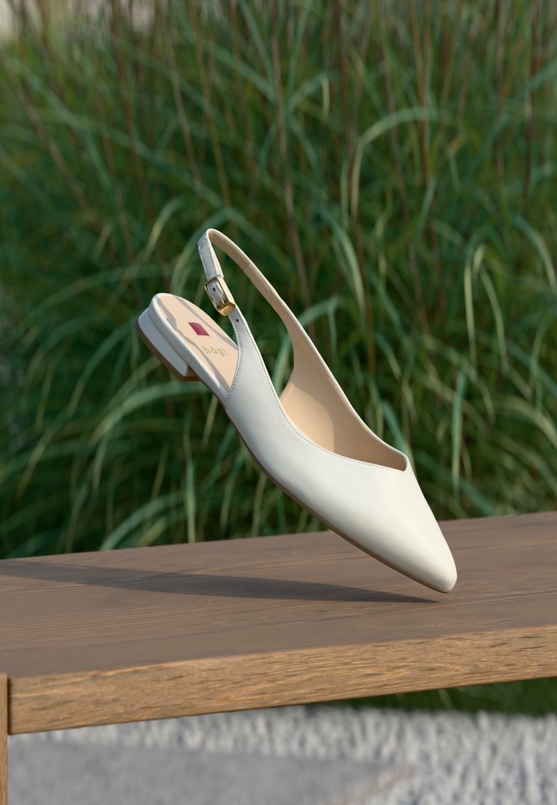 Άσπρο δερμάτινο φλατ παπούτσι slingback με μυτερή μύτη και χρυσή αγκράφα, που αιωρείται πάνω από ξύλινη επιφάνεια με πράσινα φυτά στο φόντο.