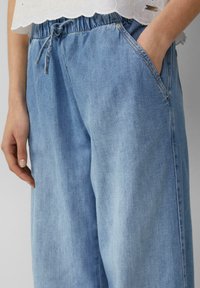 Svijetloplave traperice s elastičnom gumom u struku, vezicom i bočnim džepovima; glatka tekstura i široka kroj.