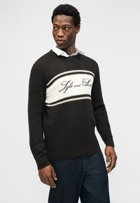 Άνδρας που φοράει μαύρο πουλόβερ με λευκή ρίγα και το κείμενο "Lyle and Scott", λευκό πουκάμισο με γιακά από κάτω και σκούρο μπλε τζιν, κοιτάζοντας προς το πλάι.