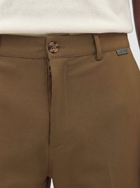 Close-up af brune bukser med delvist åben lynlås, knap er fastgjort, og et lille LUAN mærke nær taljen.