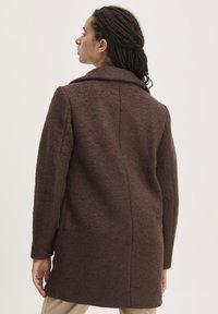 Manteau en laine texturée marron avec un large col, des manches longues et une coupe ajustée. Les détails des coutures mettent en valeur le design structuré.
