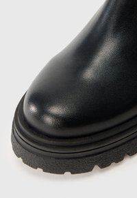 Botín de cuero negro hasta el tobillo con punta redonda, superficie texturizada y una suela de goma gruesa y resistente con dibujos visibles.
