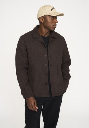 Brun tekstureret jakke med krave og forreste knapper, parret med en sort skjorte og sorte bukser. Beige kasket med sort tekst.