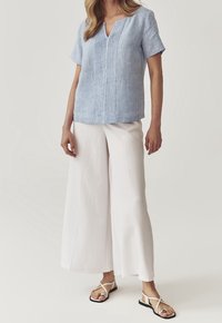 Vrouw in een lichtblauwe blouse met korte mouwen en verticale plooien, witte wijde broek en beige platte sandalen met teeringen.