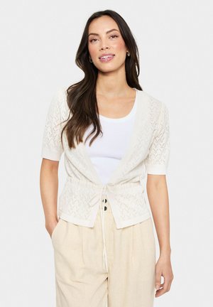 Cardigan en tricot crème avec des manches courtes, une taille à nouer et un design ouvert à motif géométrique. Associé à un top blanc.