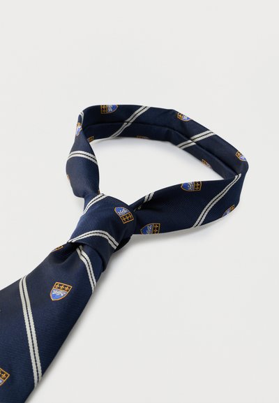 Σκούρα μπλε γραβάτα με λευκές διαγώνιες ρίγες και μικρά χρυσά και μπλε εμβλήματα ασπίδας, δεμένη με κόμπο Windsor σε λευκό φόντο.