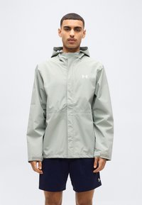 Άνδρας που φορά ανοιχτό γκρι μπουφάν με κουκούλα και σκούρο μπλε σορτς, στέκεται μπροστά σε απλό λευκό φόντο.