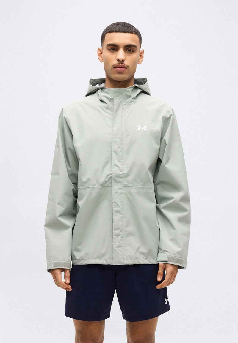 Άνδρας που φορά ανοιχτό γκρι μπουφάν με κουκούλα και σκούρο μπλε σορτς, στέκεται μπροστά σε απλό λευκό φόντο.