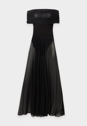 Femme portant une robe noire transparente asymétrique avec une jupe fluide, debout contre un fond clair uni.