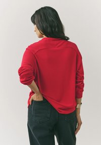 Rode pullover trui met een geribde halslijn en manchetten, met een slouchy pasvorm. Gepaard met zwarte jeans met opgestikte zakken.