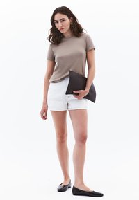 Lichtbruine top met korte mouwen, witte denim shorts, zwarte platte schoenen, houdt een donkerbruine clutch vast. Eenvoudig, getailleerd ontwerp met een gladde textuur.