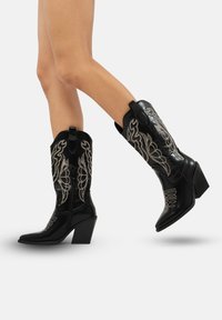 Botas de cowboy de cuero negro con punta afilada, tacón medio y elaborados bordados en plata en el caña. Cuenta con lengüetas en los laterales para facilitar su uso.