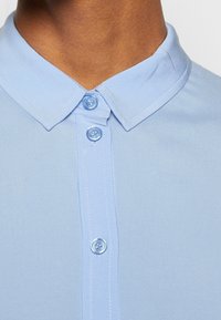Gros plan d'une personne portant une chemise bleu clair à col boutonnée jusqu'en haut avec trois boutons visibles et des coutures soignées.