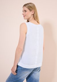 Weißes, ärmelloses Top aus Leinen mit rundem Ausschnitt und Knopfverschluss am Rücken, kombiniert mit hellblauen Jeans.