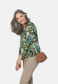 Bluse mit floralem Muster in Grün, lange Ärmel, kombiniert mit beigen Hosen und einer kleinen braunen Umhängetasche. Glattes Material und taillierte Passform.