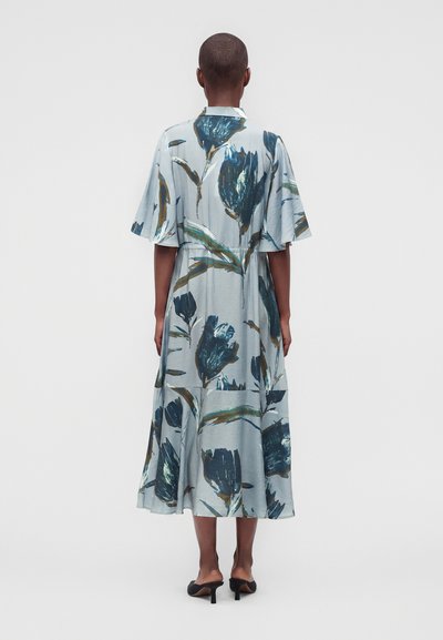 Personne portant une robe midi bleu clair avec de grands imprimés floraux bleu foncé, des manches courtes larges et des chaussures à talons noires ouvertes à l'arrière, debout de dos.