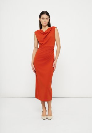Femme debout portant une robe midi orange sans manches avec un décolleté drapé et des escarpins beiges pointus, sur un fond blanc uni.