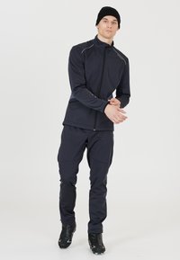 Mörkblå sportjacka och byxor för män, tillverkade av flexibelt material, med hög krage, dragkedjeslutning och reflexdetaljer. Svarta skor fulländar outfiten.