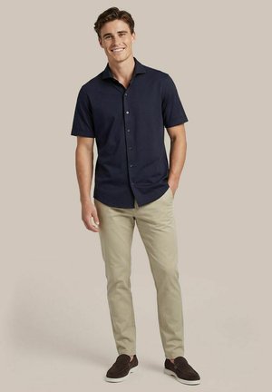 Jonge man die lacht, staat met één hand in de zak, draagt een donker marineblauw overhemd met korte mouwen en knopen, beige broek en donkerbruine schoenen.