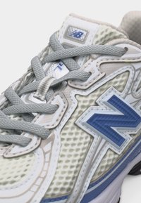 Κοντινό πλάνο αθλητικού παπουτσιού New Balance με γκρι κορδόνια, μπεζ πλέγμα στο πάνω μέρος και μεγάλο μπλε λογότυπο "N" στο πλάι.