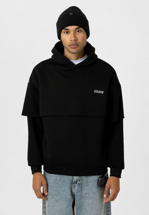 Zwarte hoodie met gelaagd ontwerp, voorzien van een logo op de voorkant. Draagt met lichtblauwe jeans en een zwarte beanie. Eenvoudige, moderne esthetiek.
