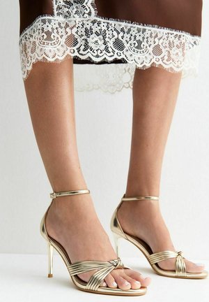 Vrouw draagt gouden sandalen met hoge hakken en enkelbandjes en een bruine jurk met witte kanten afwerking.