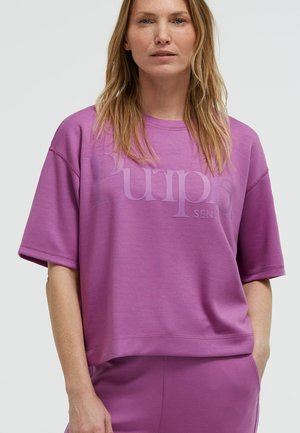 Frau mit langen blonden Haaren trägt ein locker sitzendes, lilafarbenes kurzärmeliges Oberteil und passende Hose, steht mit einer Hand in der Tasche vor schlichtem Hintergrund.