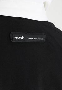 Détail du dos d'une veste noire avec une étiquette noire cousue portant l'inscription « RECCO Advanced Rescue Technology » en texte blanc près du col.
