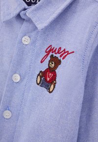 Camicia azzurra chiaro con colletto realizzata in tessuto con una trama testurizzata. Presenta bottoni bianchi e un orsetto ricamato in un maglione rosso con la scritta "guess."