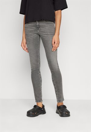Persoon die grijze skinny jeans, een zwart shirt met korte mouwen en zwarte instapklompen draagt, staand tegen een witte achtergrond.