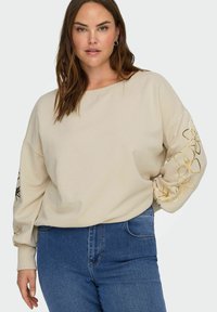 Beige-coloured collegepaita leveällä venekaula-aukkolla, tiputetut olkapäät ja pitkät hihat, joissa kultaiset kukkaompeleet vasemmassa hihassa. Puettu sinisten farkkujen kanssa.