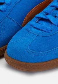 Close-up of blue suede sneakers with matching laces and brown textured rubber soles on a white surface.