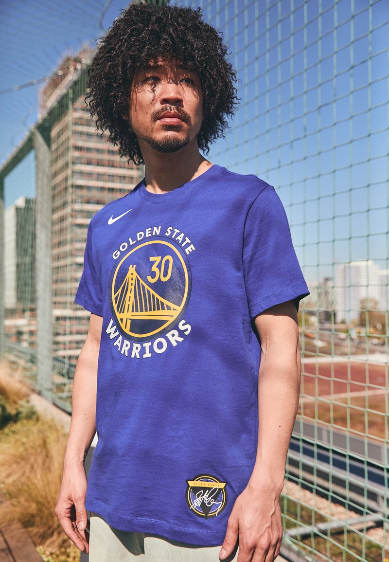 Tricou din bumbac albastru cu "Golden State Warriors" imprimat în alb și galben. Include un logo cu un pod și numărul 30 al unui jucător. Mâneci scurte.