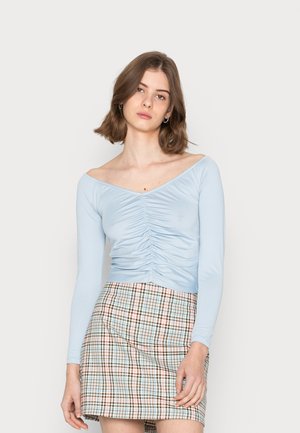 Camisola justa de manga longa azul claro com detalhes franzidos na frente, combinada com uma saia curta xadrez em cores pastel.