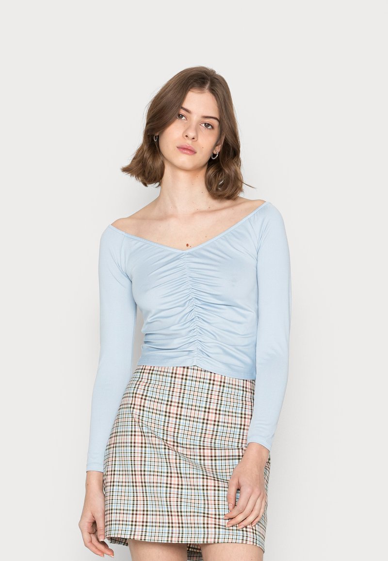 Camisola justa de manga longa azul claro com detalhes franzidos na frente, combinada com uma saia curta xadrez em cores pastel.
