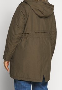 Chaqueta parka de color verde oliva con capucha, cintura elástica y aberturas laterales; presenta una tela texturizada y detalles de costura en las mangas.