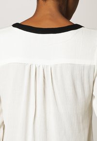 Chemise blanche texturée avec un col noir. Comprend un pli au dos pour un design supplémentaire. Le tissu a une apparence douce et légèrement froissée.