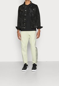 Schwarze Denimjacke, schwarzes T-Shirt, hellgrüne Jogginghosen mit seitlichen Akzenten und schwarze Sneakers mit weißen Details, die ein lässiges Outfit bilden.