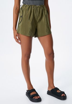 Pantalones cortos verde olivo con cintura elástica y cordón, con ribetes negros y bolsillos laterales. Combinados con sandalias negras.