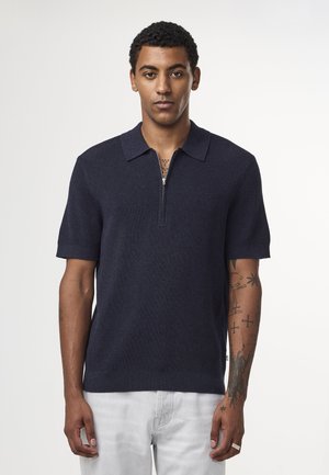 Marineblauwe polo met korte mouwen van gebreide stof, voorzien van een ritssluiting bij de kraag, een geribbelde textuur en subtiele details op de mouw, gecombineerd met lichte jeans.