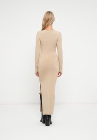Langermet, tettsittende kjole i lys beige stoff, med en sidesplitte og glatt tekstur, kombinert med knehøye sorte støvler.