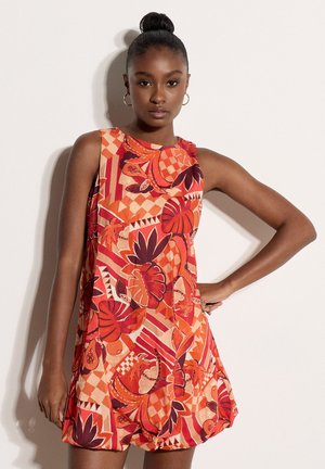 Femme avec les cheveux en chignon, portant une robe sans manches orange et rouge à motifs de feuilles tropicales et dessins géométriques, debout devant un fond clair.