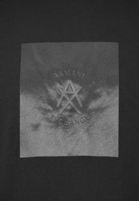 Tecido preto com um quadrado cinza apresentando um logotipo em relevo da Armani Exchange e texto centralizado dentro do quadrado.