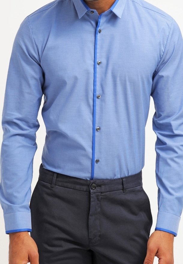 Homme portant une chemise bleu clair boutonnée avec une bordure bleu plus foncé aux poignets et sur la patte de boutons, associée à un pantalon gris foncé.