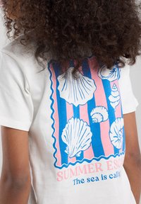 White t-shirt featuring a colorful shell print in blue and pink, with scalloped edges and the text “SUMMER ESCAPE” and “The sea is calling.”