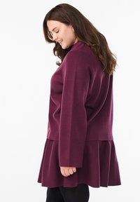 Žena s dlouhými hnědými vlasy, oblečená v odpovídajícím svetr a sukni v barvě švestky, usmívající se a dívající se dolů na bílém pozadí.