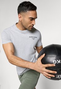 Gray short-sleeve t-shirt with a black logo, combined with green shorts. The person holds a black medicine ball. Focus on fitness attire.