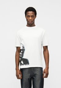 Άνδρας που στέκεται και φοράει λευκό T-shirt με μαύρο γραφικό σχεδιάγραμμα ενός προσώπου που φοράει γυαλιά ηλίου στην κάτω αριστερή πλευρά.