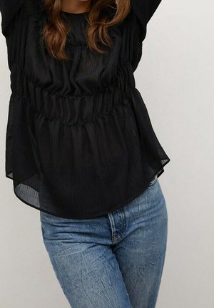 Femme portant un chemisier noir ample et texturé avec une taille froncée et un jean bleu en denim sur un fond uni.